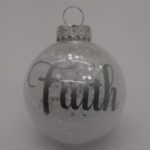 May include: A clear glass Christmas ornament with silver glitter inside and the word "Faith" written in black lettering.