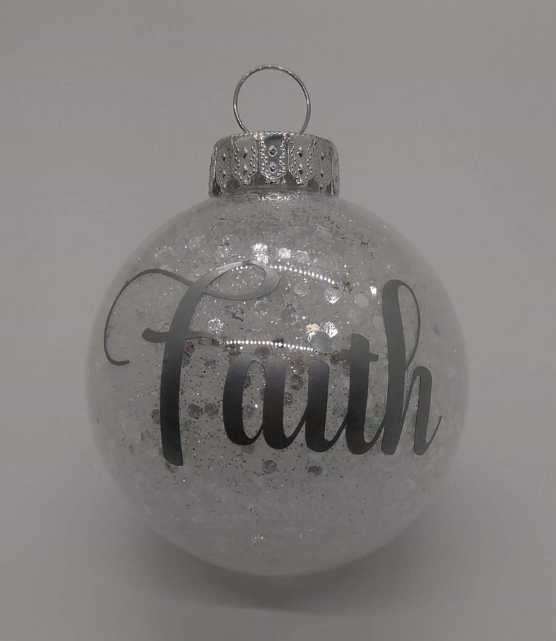 May include: A clear glass Christmas ornament with silver glitter inside and the word "Faith" written in black lettering.