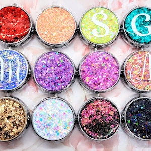 Può includere: Dodici contenitori rotondi con bordo argentato, con glitter di diversi colori e una lettera bianca su ogni coperchio. Le lettere sono M, S, G, A e un design di glitter.