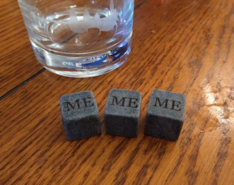 May include: Three gray whiskey stones with the word "ME" engraved on each, arranged on a wooden surface. A clear glass with a city skyline design and the word "Friends" is in the background.