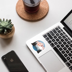 May include: A sticker featuring a mountain landscape with a blue sky, snow-capped peaks, and pine trees. The sticker is on a silver laptop.
