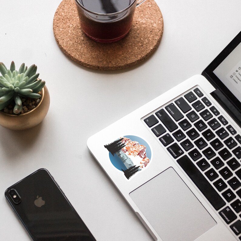 May include: A sticker featuring a mountain landscape with a blue sky, snow-capped peaks, and pine trees. The sticker is on a silver laptop.