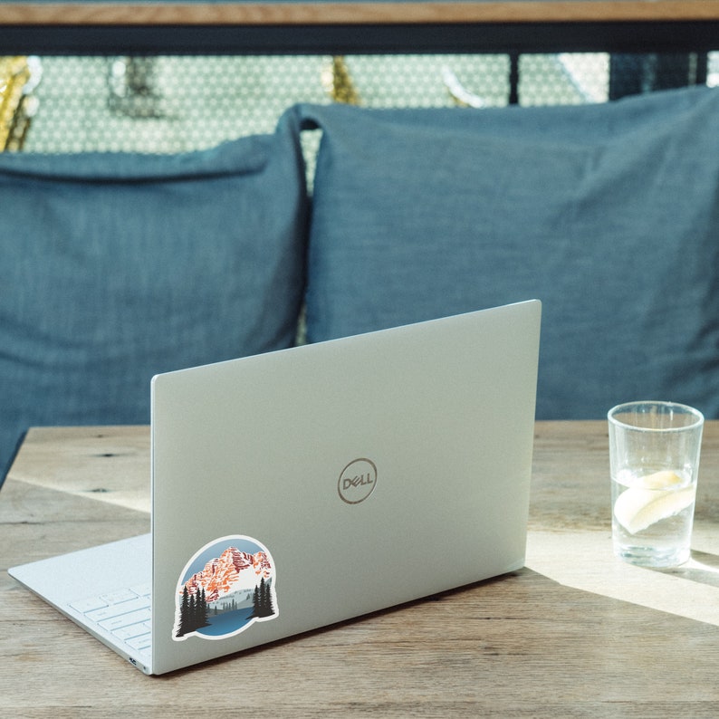 May include: A silver laptop computer with a sticker of a mountain range with a lake in front of it. The sticker is on the back of the laptop.