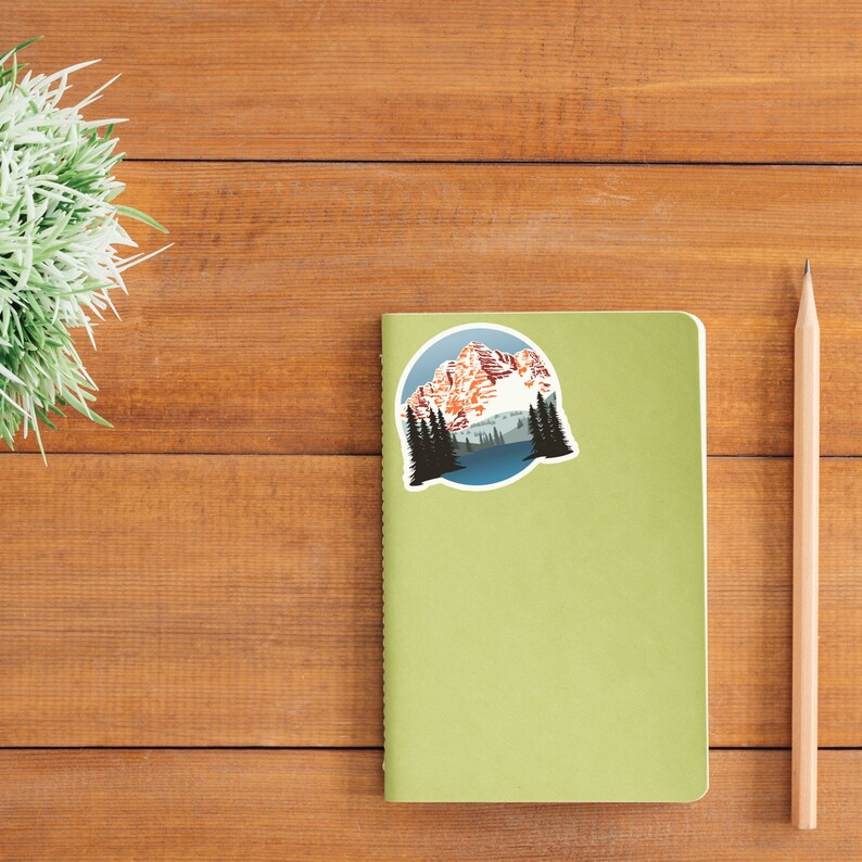 May include: A green notebook with a round sticker featuring a mountain range with snow-capped peaks and a lake in the foreground. The sticker is surrounded by a blue circle.