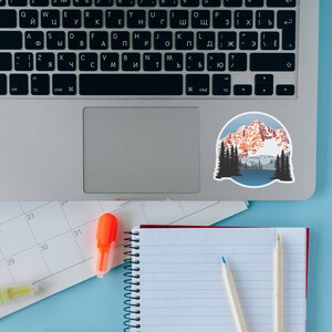 May include: A laptop computer with a sticker of a mountain range with a blue lake in the foreground. The laptop is on a blue surface with a calendar, a notebook, a highlighter, and two pencils.