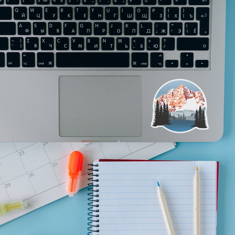 May include: A laptop computer with a sticker of a mountain range with a blue lake in the foreground. The laptop is on a blue surface with a calendar, a notebook, a highlighter, and two pencils.