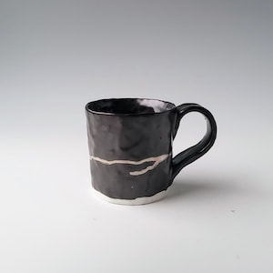 May include: A black ceramic mug with a white glaze and a gold line design. The mug has a rounded handle.