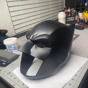 May include: A black, moulded mask with pointed ears, reminiscent of a superhero's cowl. The mask features a defined brow line, eye openings, and a prominent, angular nose piece. The surface appears smooth and matte.