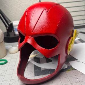 May include: A vibrant red Flash helmet with a distinctive lightning bolt design on the forehead. The helmet features eye openings and yellow side wings, showcasing a superhero theme. The design is sleek and detailed, perfect for cosplay or display.
