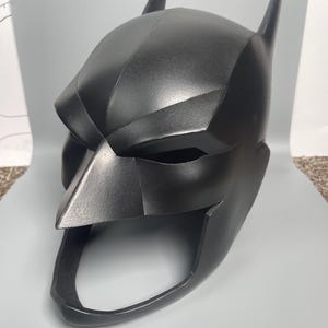 May include: A black Batman cowl with pointed ears. The mask has a textured surface and a defined facial structure, including a prominent nose and eye openings. The cowl is designed to fit the head and neck.
