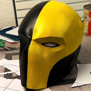 Może przedstawiać: Ręcznie wykonany, dwukolorowy hełm w kolorze czarnym i żółtym, prawdopodobnie do cosplayu. Hełm jest podzielony pionowo, z jednej strony czarny, a z drugiej żółty. Otwór na oczy ma siatkę. Artykuł kostiumowy.