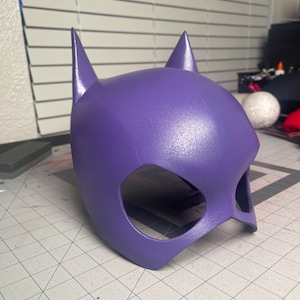 May include: A purple superhero mask with pointed ears and eye openings. The mask is made of a smooth material and is designed to fit the head. The mask is a solid, vibrant purple color.