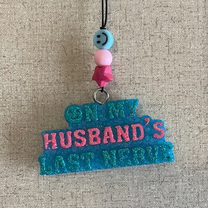 May include: A turquoise blue necklace with a pink star charm and a blue smiley face bead. The charm has the text "On My Husband's Last Nerve" in pink glitter.