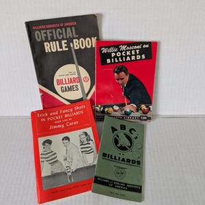 May include: A collection of vintage billiard books. Titles include "Official Rule Book," "Willie Mosconi on Pocket Billiards," "Trick and Fancy Shots in Pocket Billiards," and "ABC's of Billiards." The books are in various colors, including red, black, and green.