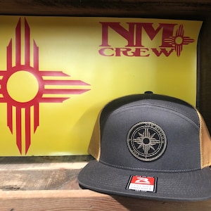 May include: A black and gold trucker hat with a compass design and the text "New Mexico" on the front. The hat is sitting in front of a yellow sign with the New Mexico state symbol and the text "NM Crew".