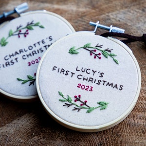 May include: Two embroidered Christmas ornaments with a holly design. The first ornament says "Charlotte's First Christmas 2022" and the second ornament says "Lucy's First Christmas 2023".