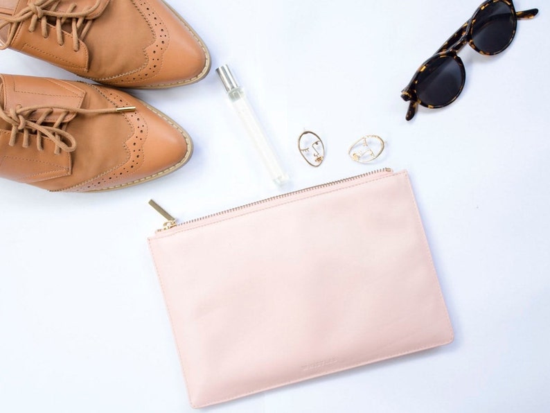 May include: A light pink clutch bag with a gold zip, brown leather shoes, gold earrings, and brown tortoiseshell sunglasses on a white background.