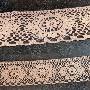 May include: Close-up of two strips of peach-colored lace. The lace features an openwork design with floral and geometric patterns. The edges are scalloped, and the lace appears to be made of a soft, delicate material. The lace is likely used for sewing and crafting.