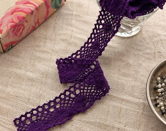 May include: A deep purple crochet lace trim featuring a repeating circular pattern with scalloped edges. The lace is arranged in a loose, flowing strip, showcasing the intricate openwork design and the vibrant, saturated color of the thread.