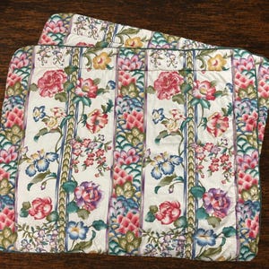 May include: A square quilted pillow sham featuring a white background with vertical stripes of green braided patterns and floral motifs in shades of pink, red, purple, yellow, and blue. The edges are finished with a dark green piped trim.