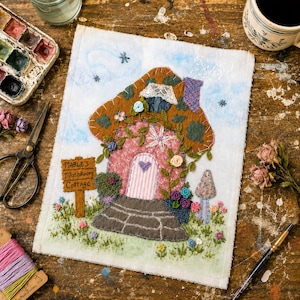 Op de afbeelding: Een stoffen wandkleed met een geborduurd paddenstoelhuisje met een bruine hoed, een roze gestreepte deur en een bordje met de tekst Mable's Mushroom Cottage. De scène bevat gestikte bloemen, klimplanten en een stenen pad op een blauwe achtergrond.