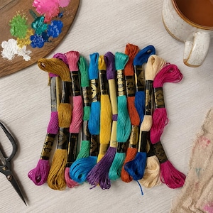 May include: A collection of twelve multi-colored embroidery floss skeins with black paper labels marked DMC 25. The threads feature vibrant shades of gold, pink, green, blue, purple, orange, and cream, arranged in a vertical row on a light surface.