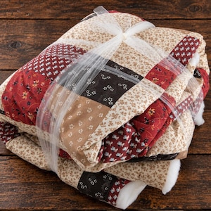 May include: A folded patchwork quilt featuring a variety of square fabric panels in shades of deep red, dark brown, and cream. The quilt is tied with a white tulle ribbon and displays floral and geometric patterns across the quilted surface.