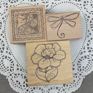 May include: Three wooden stamps with black ink designs. One features a butterfly in a decorative frame, another a bow, and the third a teddy bear sitting in a flower. These stamps are ideal for scrapbooking and crafting.