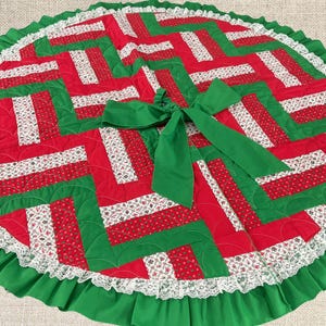 May include: A circular quilted tree skirt featuring a geometric pattern of red, green, and white floral fabric strips. The skirt is finished with a green fabric ruffle, a white lace trim along the edge, and a large green fabric bow at the center.