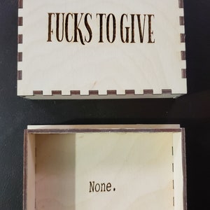 May include: A wooden box with the text "F*CKS TO GIVE" on the lid and the word "None." inside. The box is made of light wood with a dark brown border.