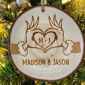 May include: A wooden Christmas ornament with a heart shape formed by two hands. The hands are holding a heart with the initials "M+J" inside. The ornament is inscribed with the names "MADISON & JASON".