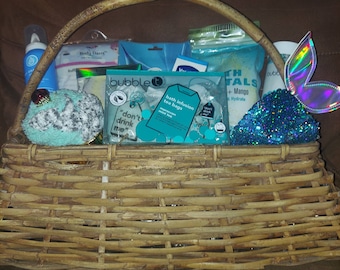 Può includere: Un cesto di vimini pieno di prodotti per il bagno e il corpo, tra cui sali da bagno, bombe da bagno, bustine di tè e un panno da bagno. Il cesto include anche una coda di sirena blu e verde e una bottiglia di bagnoschiuma.