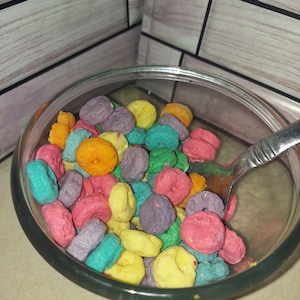 May include: A clear glass bowl filled with colorful, ring-shaped cereal. The cereal pieces are in various colors, including pink, purple, yellow, orange, and teal. A silver spoon rests inside the bowl, ready to eat. The background is a light-colored surface.