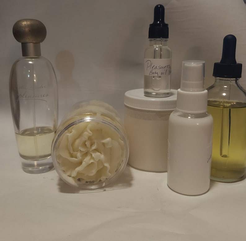 Puede incluir: Una colecci&oacute;n de productos para el ba&ntilde;o y el cuerpo que incluye una botella de vidrio transparente de aceite corporal etiquetada como "Pleasures Body Oil 1 oz", un tarro blanco de manteca corporal, una botella de vidrio transparente de perfume, una botella blanca de spray corporal y una botella de vidrio transparente de aceite.