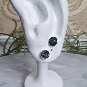 Peut inclure: Une paire de boucles d'oreilles en argent avec des accents de pierres précieuses vertes et violettes. Les boucles d'oreilles sont présentées sur un modèle d'oreille blanc.