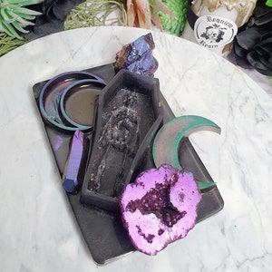 May include: Black resin tray with a coffin, crescent moon, and crystal shapes. The tray has a iridescent finish and features a skeleton inside the coffin.