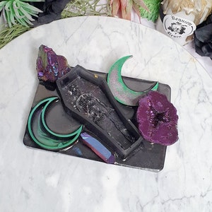 May include: A black resin tray with a coffin-shaped mold, two green crescent moons, a purple crystal, and a iridescent crystal. The tray is on a white marble surface.  The text "Requiem Resin" is visible on a small tag.