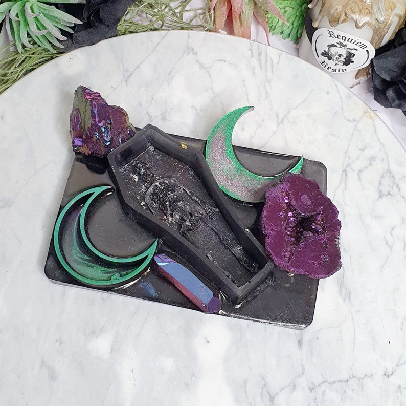 May include: A black resin tray with a coffin-shaped mold, two green crescent moons, a purple crystal, and a iridescent crystal. The tray is on a white marble surface.  The text "Requiem Resin" is visible on a small tag.