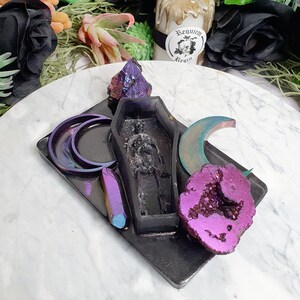 May include: A black tray with a variety of resin crafts, including a coffin-shaped mold, a crescent moon, a crystal, and a purple geode. The tray is on a white marble surface. The text "Requiem Resin" is visible on a sticker.