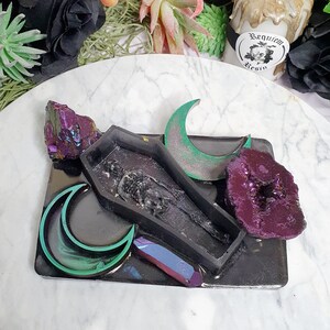 May include: Black tray with a coffin-shaped mold, two crescent moon shapes, a purple crystal, and a larger purple crystal with a hollow center. The tray has a black background with a glittery finish. The text "Requiem Resin" is visible on a sticker.