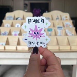 Può includere: Un adesivo con un personaggio di foglia di cannabis viola con le parole "Breathe in" e "Breathe out". L'adesivo ha uno sfondo bianco con accenti verdi e viola. L'adesivo è tenuto davanti a un espositore in legno.