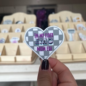 Op de afbeelding: Een hartvormige sticker met een geruit patroon in zwart en wit. De sticker heeft roze en paarse tekst die "In My Elder Emo Mom Era" luidt.