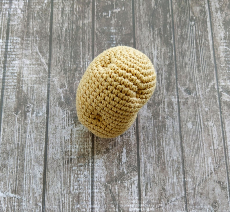 May include: A crocheted, light brown, potato-shaped toy. The toy is sitting on a wooden surface.