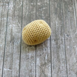 May include: A crocheted potato toy in a light brown color. The potato is sitting on a wooden surface.