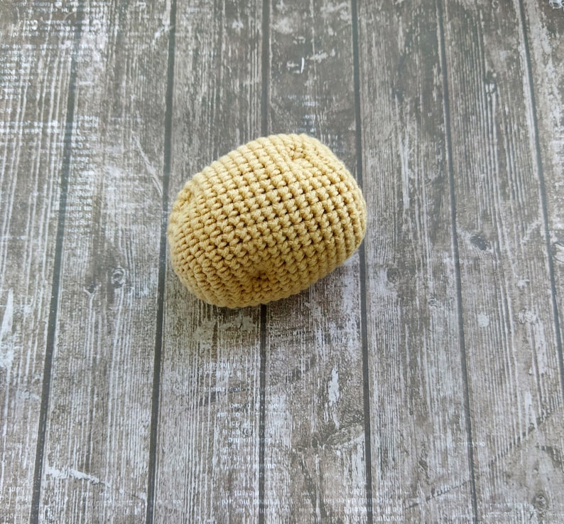May include: A crocheted potato toy in a light brown color. The potato is sitting on a wooden surface.
