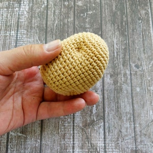 May include: A hand holding a crocheted ball in a light beige color. The ball is made of yarn and has a textured surface.