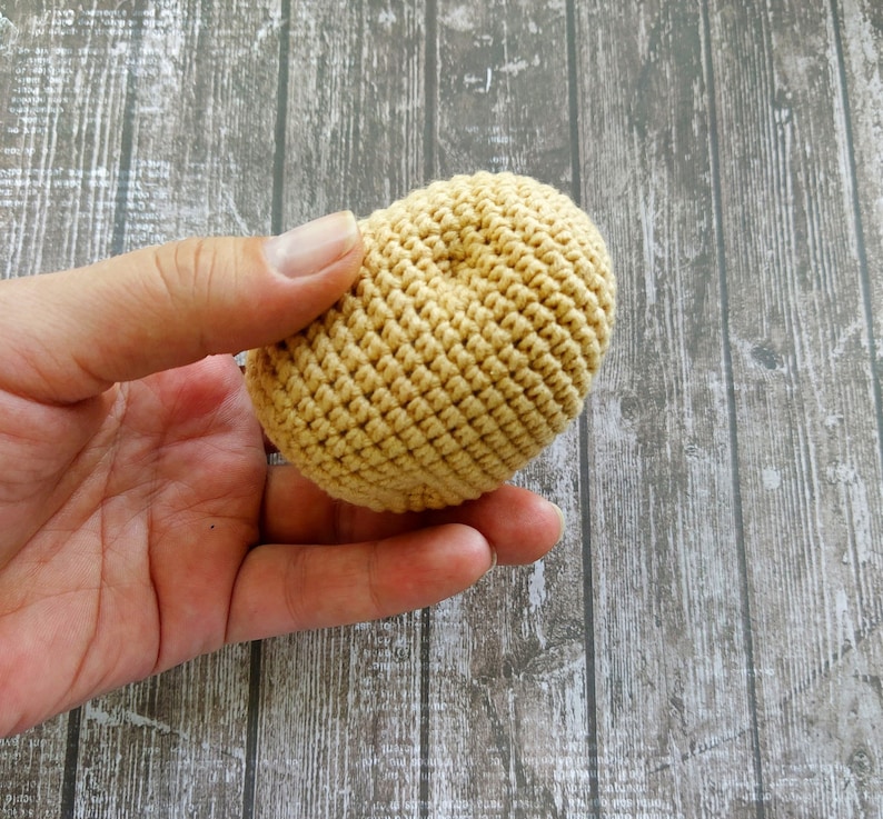 May include: A hand holding a crocheted ball in a light beige color. The ball is made of yarn and has a textured surface.