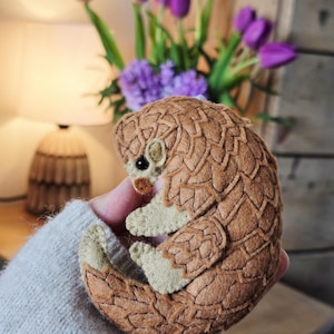 May include: A handmade felt pangolin figurine in shades of brown and beige. The pangolin is curled up, showcasing its textured scales and small, detailed features. The text "ArtandcraftMadalinaV" is visible.