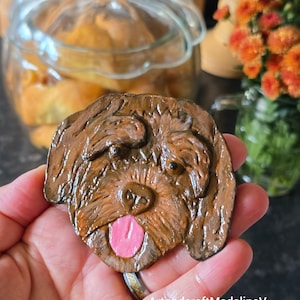 May include: A brown, handmade dog-shaped ornament with a pink tongue. The ornament is held in a hand, with the text "ArtandcraftMadalinaV" visible. The background includes a glass jar and orange flowers.