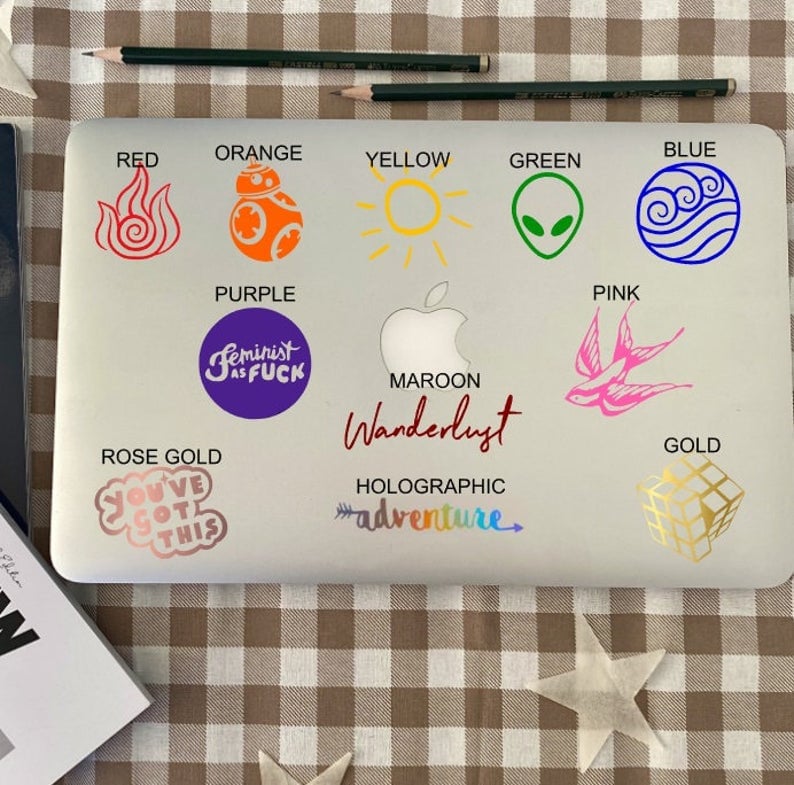 Pu&ograve; includere: Un computer portatile con vari adesivi colorati sul coperchio. Gli adesivi includono un sole, un personaggio di Star Wars, un simbolo di fuoco, un simbolo di acqua, un alieno, un uccello, un cubo, uno slogan femminista e la parola "Wanderlust".
